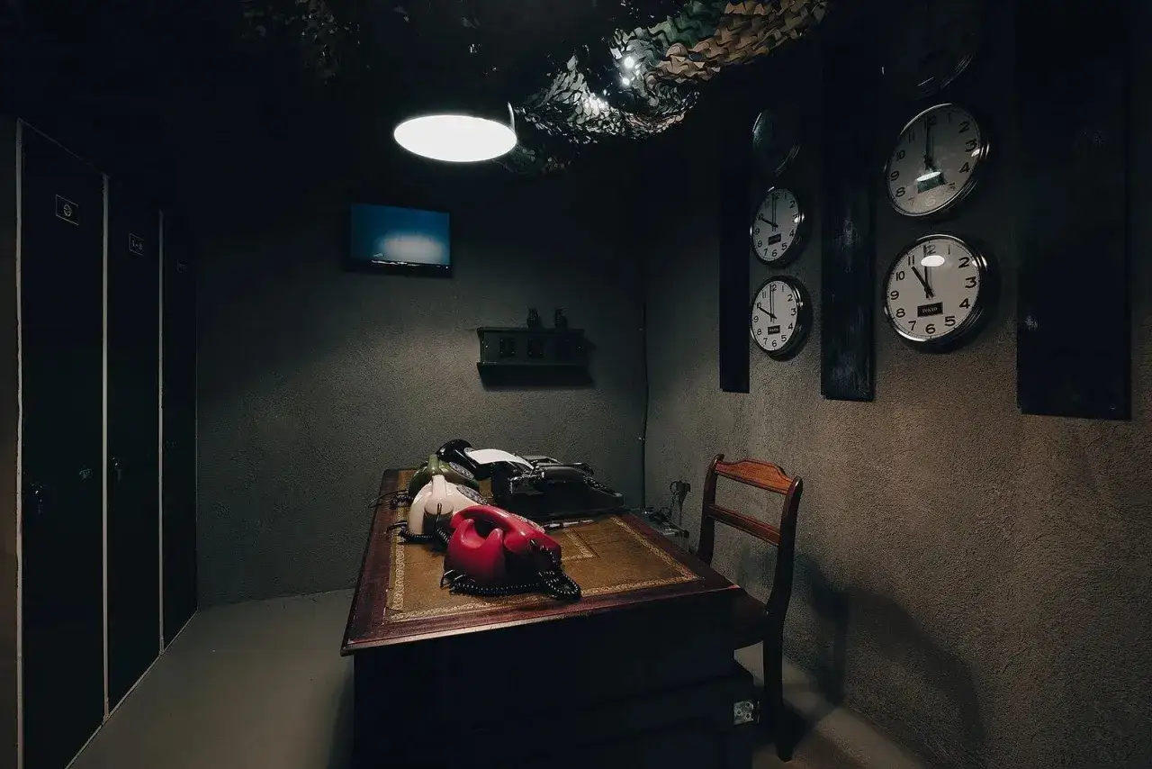 Mörkt rum med skrivbord, gamla telefoner, skrivmaskin och flera klockor på väggen. Perfekt för en spännande escape room hemma-upplevelse.