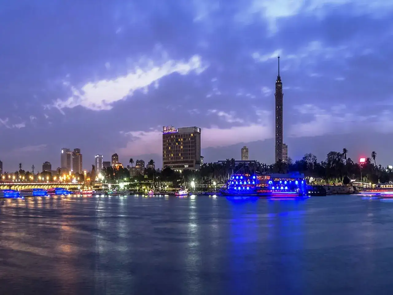 Ein beleuchtetes Schiff mit blauen Lichtern gleitet &uuml;ber den Nil. Im Hintergrund erstreckt sich die Skyline von Kairo bei Nacht, gekr&ouml;nt vom Cairo Tower.