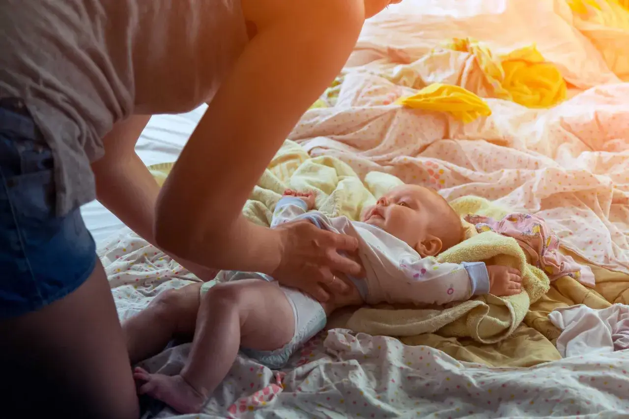 Mama delikatnie dotyka brzuszka niemowlaka chłopca, kt&oacute;ry leży na kolorowej pościeli. Może to być kontrola po diagnozie przepukliny u niemowlaka chłopca.
