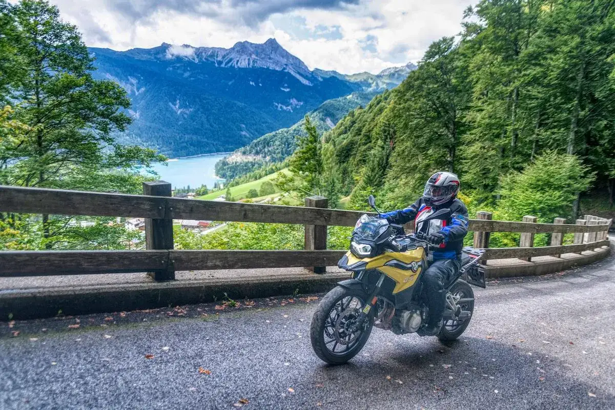 Motociclista in sella a una moto gialla su una strada panoramica con lago e montagne sullo sfondo. Un vero passo pura di avventura.