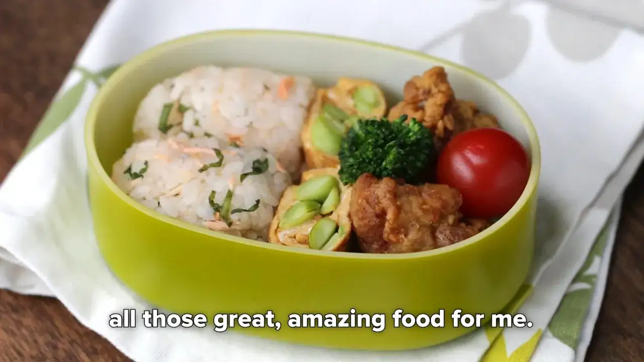 A vibrant green bento box filled with a delicious tamagoyaki bento, featuring rice balls, fried chicken, broccoli, and a cherry tomato.