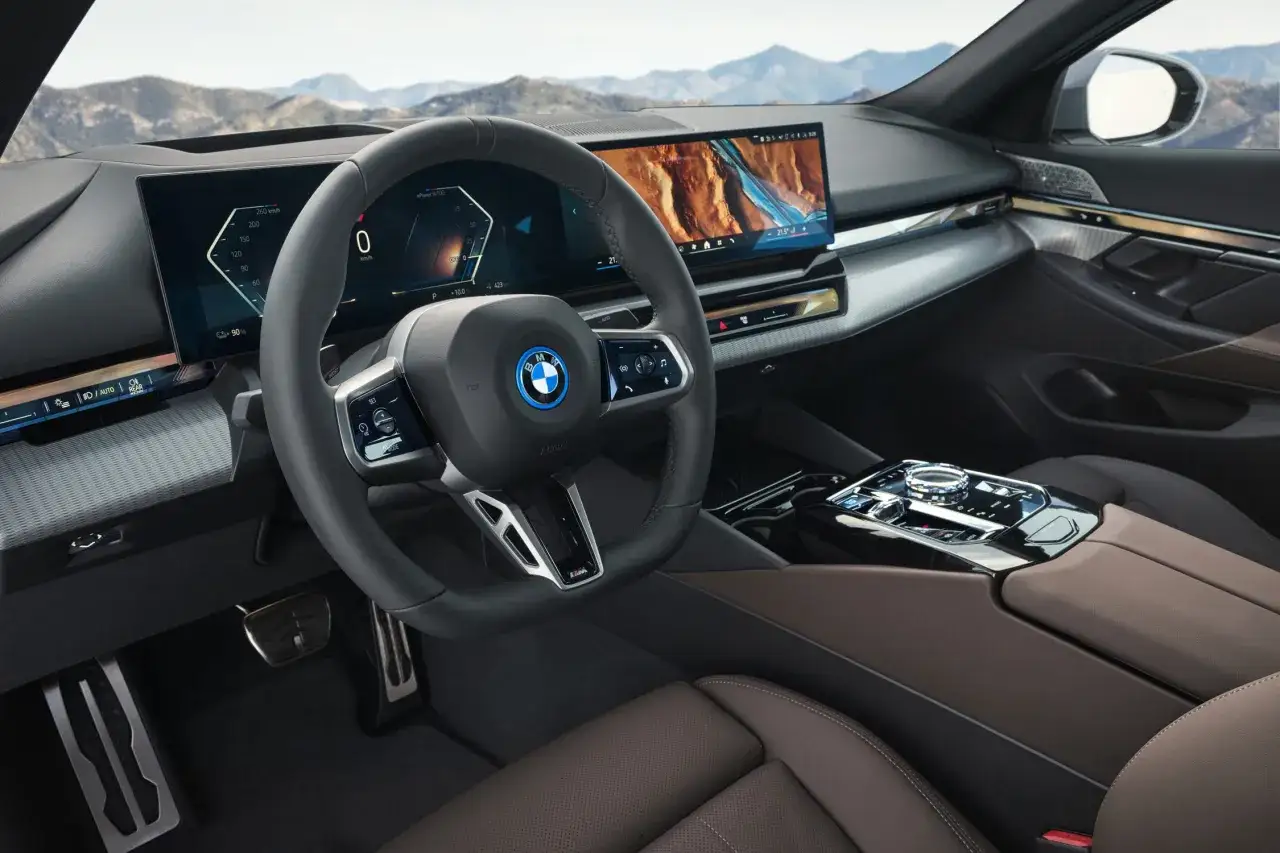 Wnętrze BMW i5 z dużymi ekranami, skórzaną tapicerką i nowoczesnym panelem sterowania. Widok na górzysty krajobraz przez przednią szybę.
