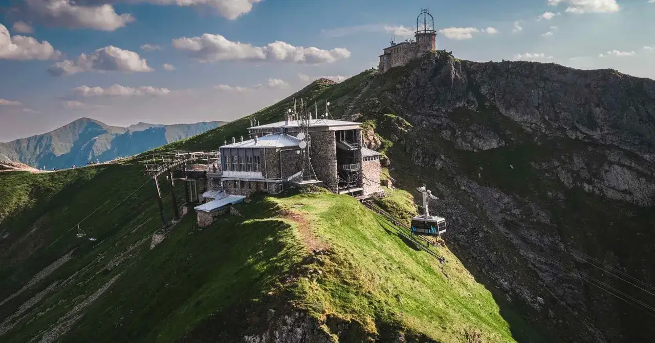 Szlak na Kasprowy Wierch z Kuźnic prowadzi przez zielone zbocza, gdzie widać budynki i kolejkę linową.