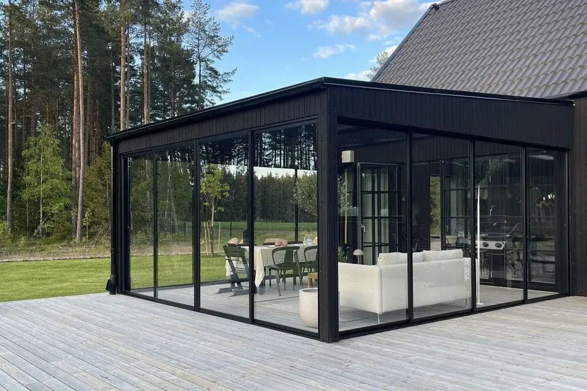 En modern inglasad altan med svarta ramar, möblerad med soffa och matgrupp. Perfekt för att njuta av naturen året runt.