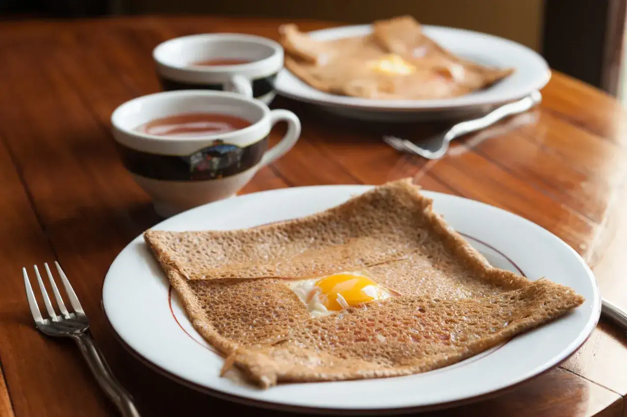 Une galette de sarrasin, spécialité de la **crêpe Bretagne**, avec un œuf au centre, servie sur une assiette blanche avec une tasse de thé.