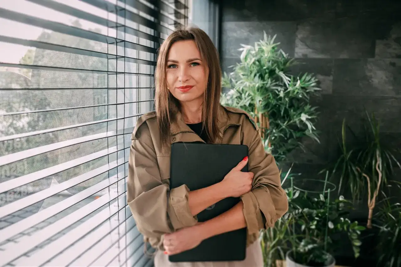 Kobieta w beżowej kurtce trzyma laptopa, stoi przy oknie z żaluzjami i roślinami.