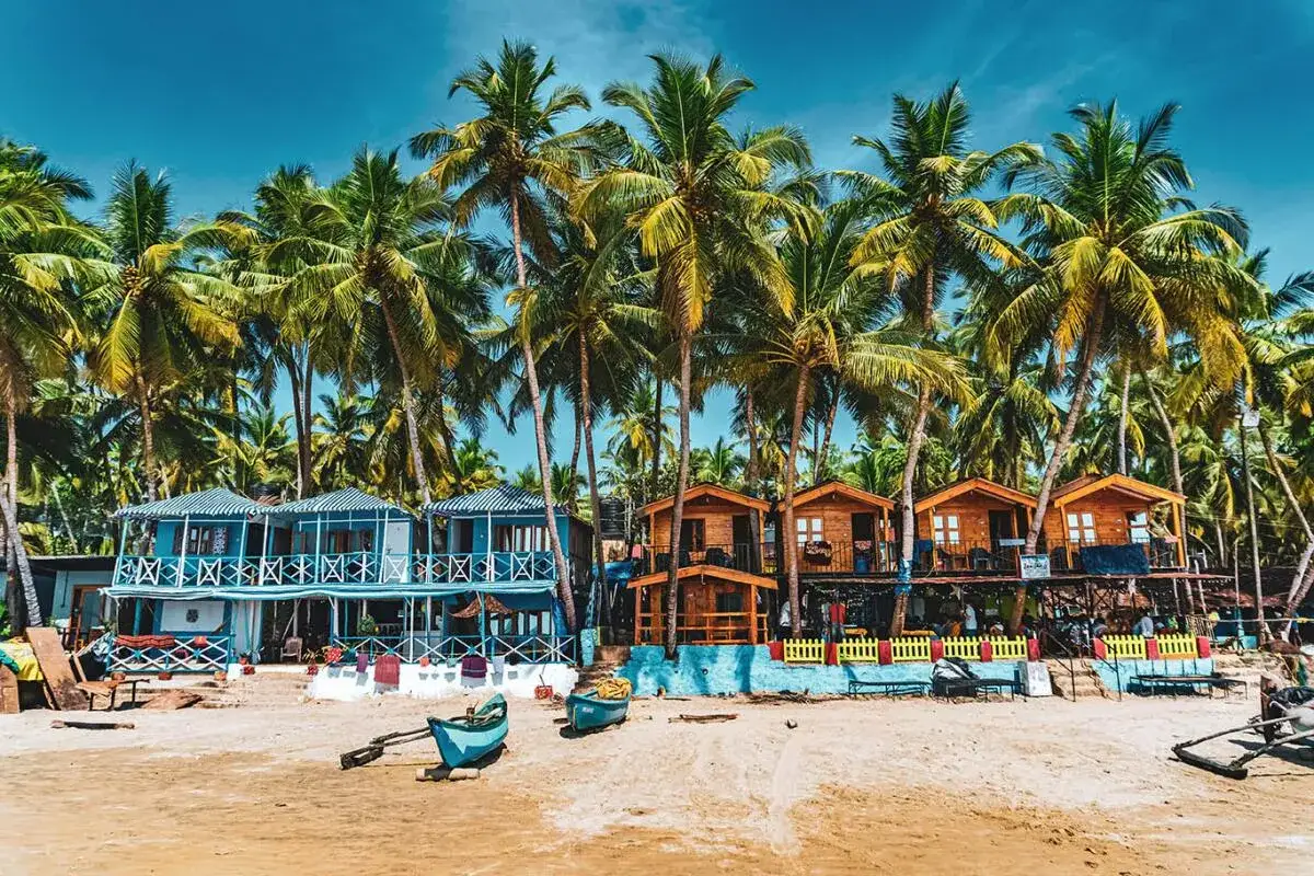 Strandbungalows och bl&aring; hus under vajande palmer vid en strand i Goa, Indien. En dr&ouml;m f&ouml;r alla som s&ouml;ker en avkopplande semester.