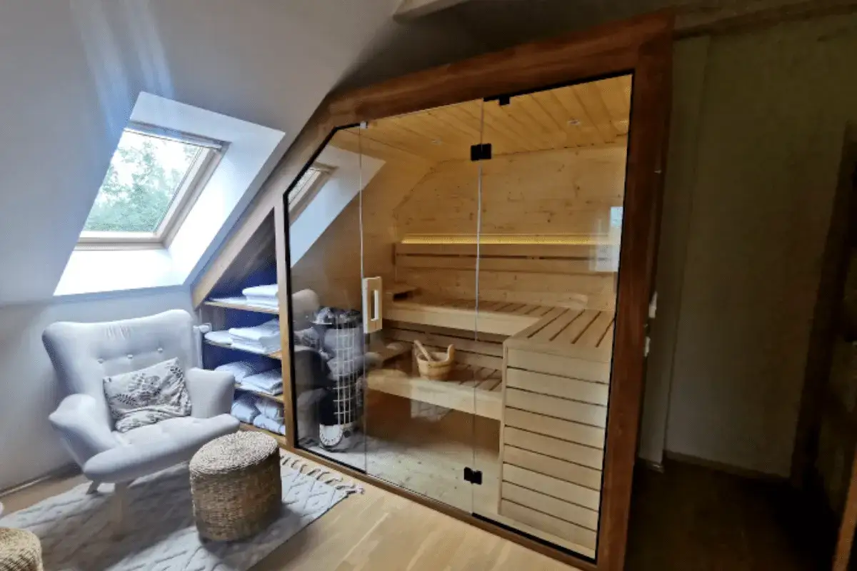 Przytulna sauna na poddaszu z przeszklonymi drzwiami. Obok wygodny fotel i półki z ręcznikami. Idealne miejsce na relaks.