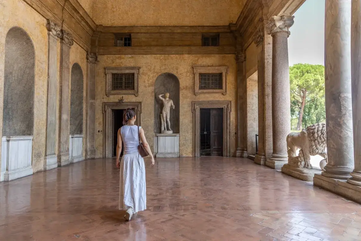 Une femme en robe blanche marche dans la cour de la Villa M&eacute;dicis, admirant une statue antique et un lion de pierre.
