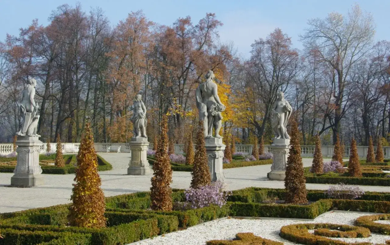 Kamienne rzeźby w ogrodzie w stylu francuskim, otoczone jesiennymi drzewami i geometrycznymi żywopłotami.