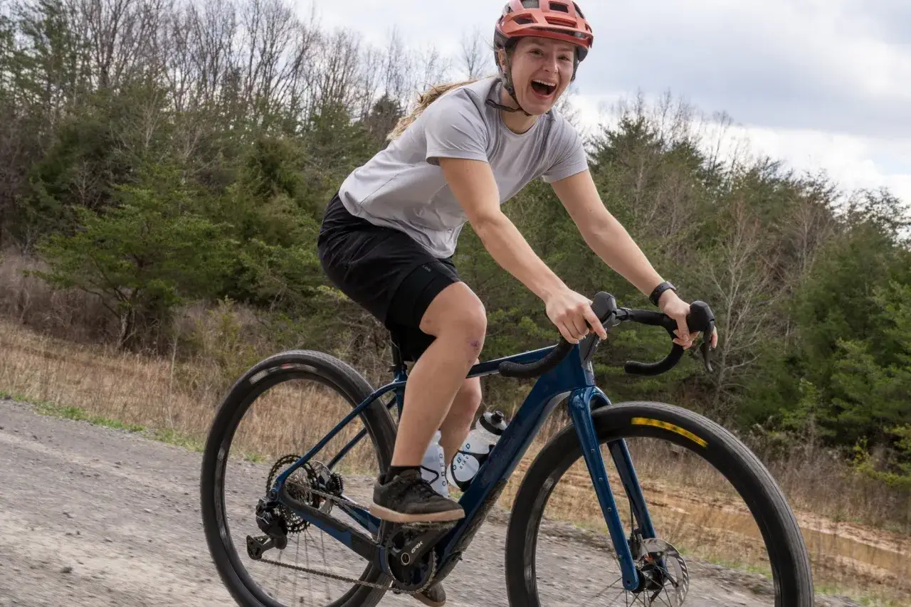 A woman with a helmet and a wide smile enjoys a gravel road, ready for long rides through the trees.
