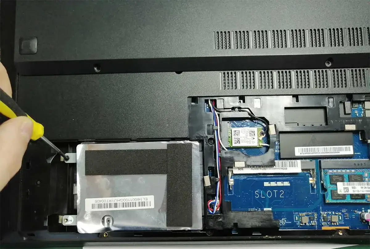 Ręka z żółto-czarnym śrubokrętem odkręca dysk twardy w laptopie. Widać moduł Wi-Fi i slot 2. Pamiętaj o ESD.