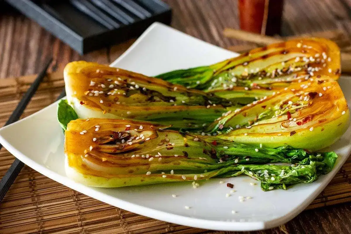 Drei Hälften von gegrilltem Pak Choi, glasiert und mit Sesam bestreut, auf einem weißen Teller.