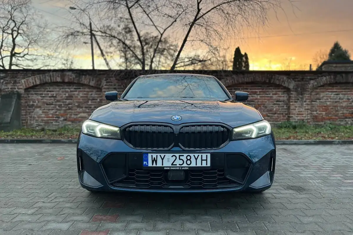 Eleganckie BMW 330e w kolorze granatowym, z włączonymi światłami, zaparkowane przed ceglanym murem o zachodzie słońca.
