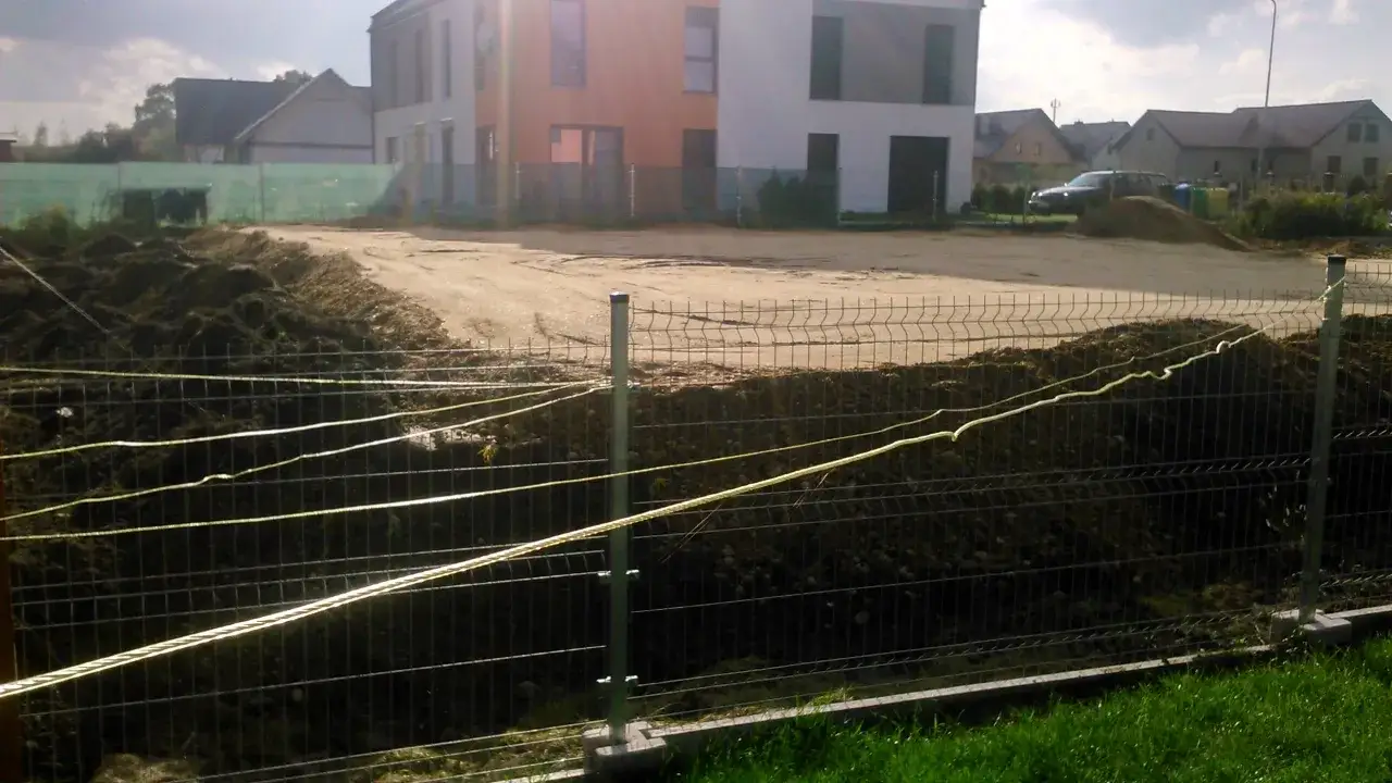 Budowa domu w toku. Koparka usypała ziemię, tworząc wał. Czy sąsiad może podnieść działkę? Płot ogrodzeniowy z żółtymi linkami.