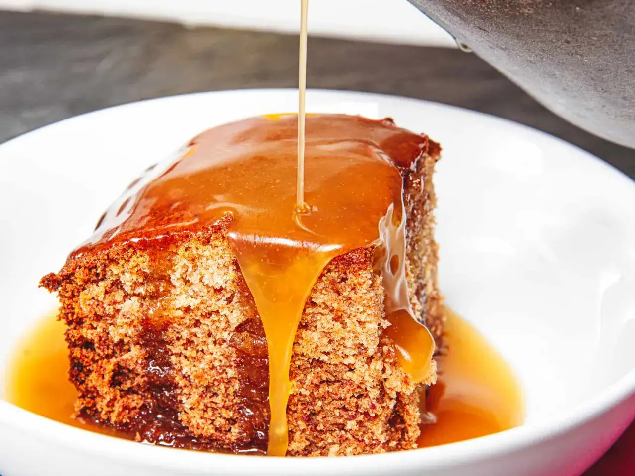 En bit saftig sticky toffee pudding dränks i varm kolasås i en vit skål.