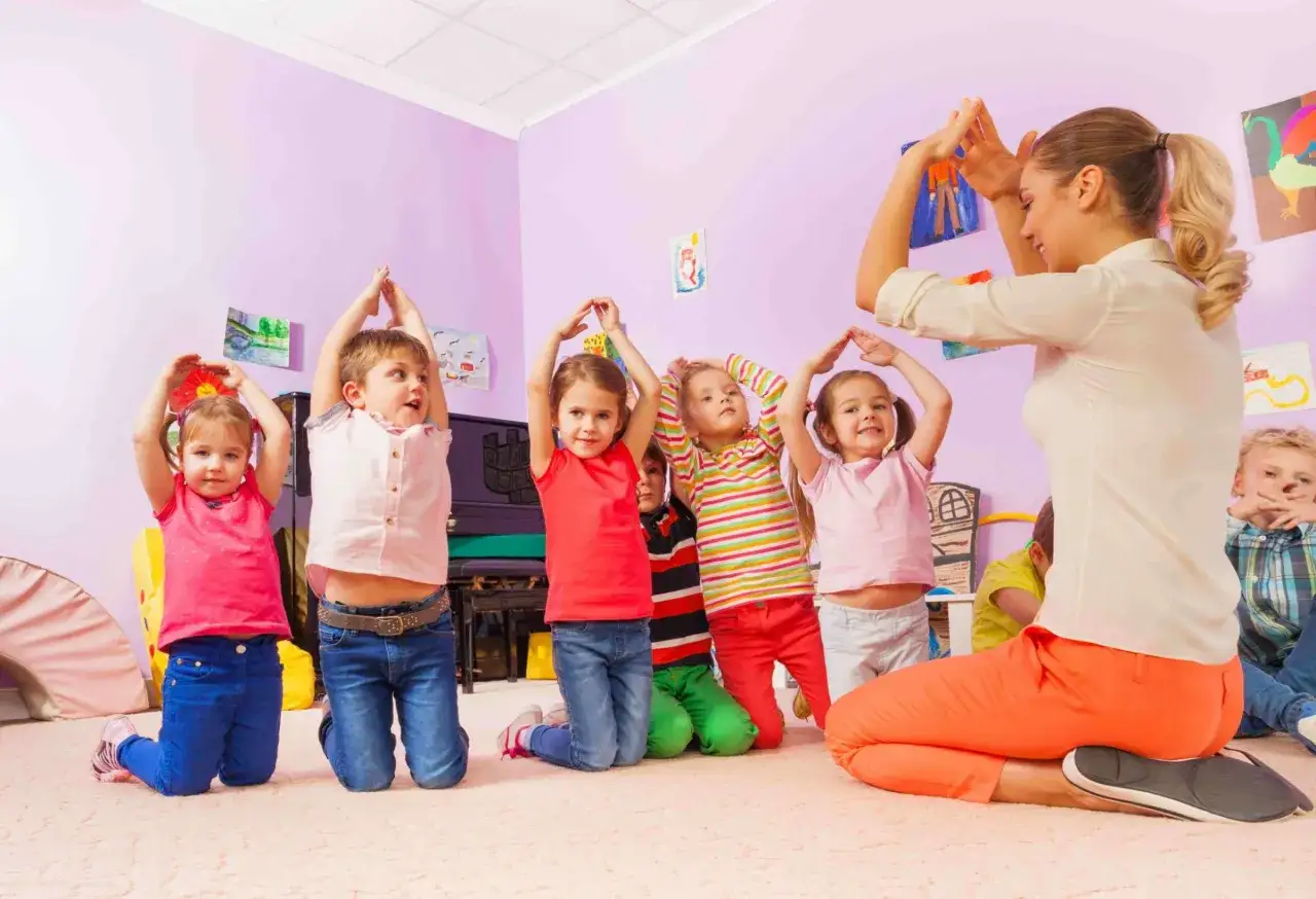 Niños y maestra participan en juegos en grupo sentados, levantando las manos en un aula colorida.