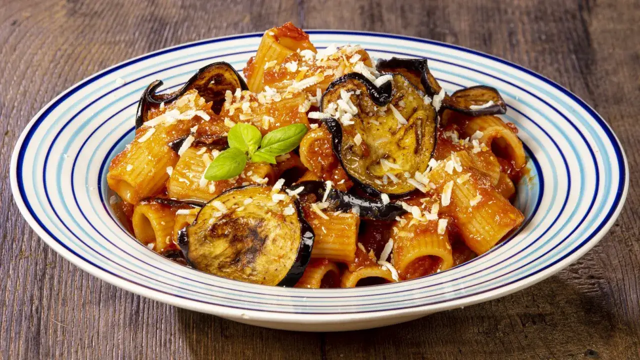 Piatto di pasta alla norma ricetta originale con melanzane fritte, pomodoro e ricotta salata grattugiata, guarnito con basilico fresco.