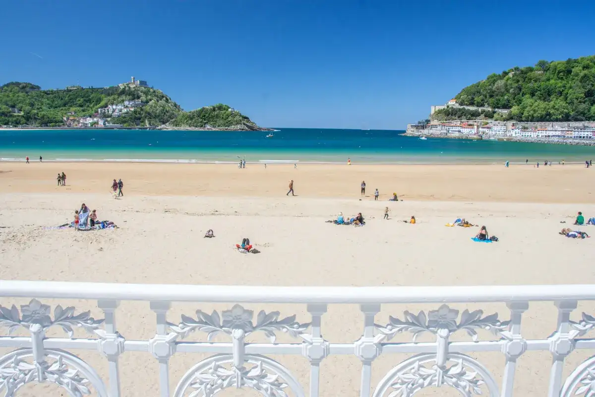 Słoneczna plaża w Donostia San Sebastian, z turkusową wodą i zielonymi wzgórzami w tle. Ludzie odpoczywają na piasku, ciesząc się pięknym dniem.