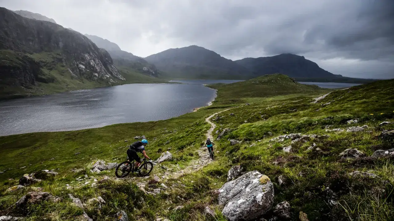 Two cyclists navigate a rocky trail with a vast lake and dramatic mountains behind them, capturing the essence of Scotland bike tours.