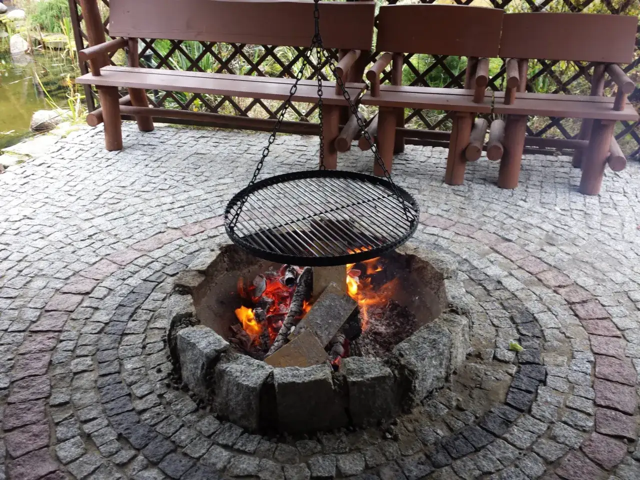 Ognisko z kostki brukowej, gotowe do rozpalenia. W tle drewniane ławki i oczko wodne.