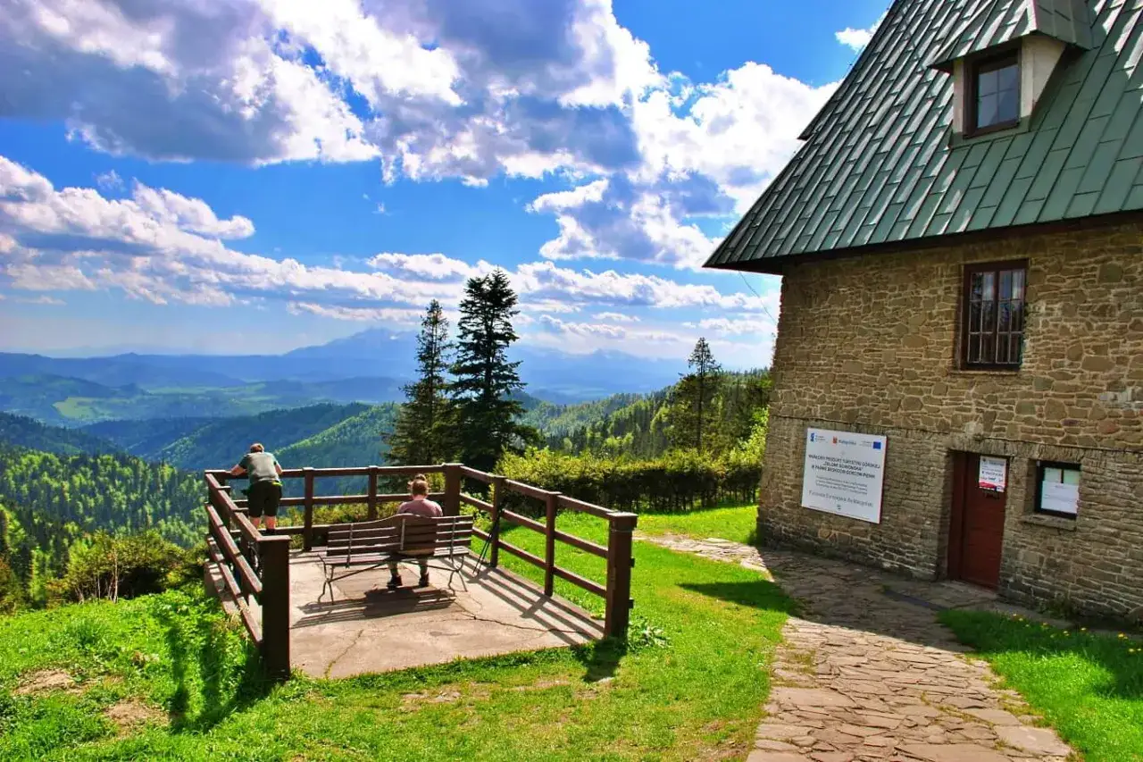 Widok na malownicze **beskid niski szczyty** z tarasu widokowego przy kamiennym schronisku. Dwie osoby odpoczywają na ławce, podziwiając panoramę.