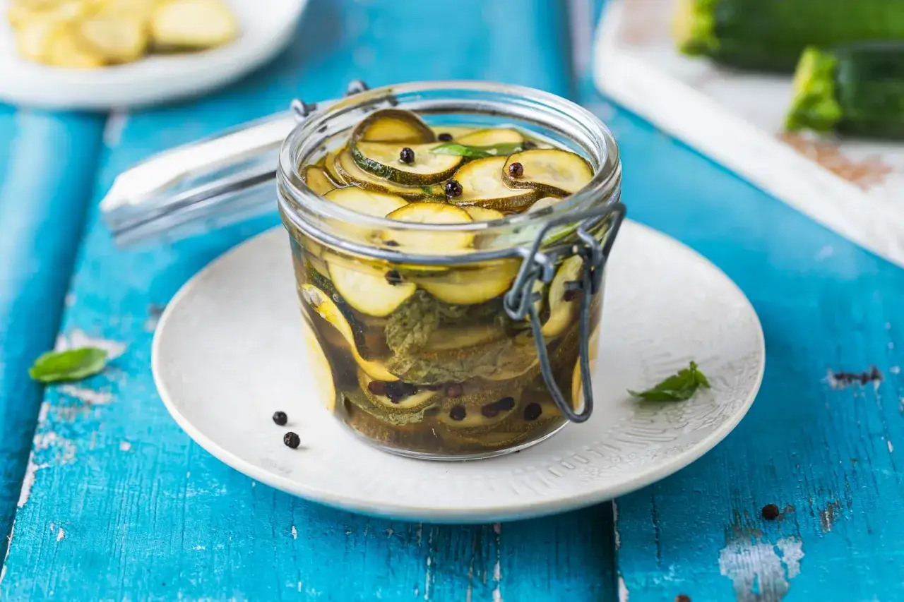 Barattolo di zucchine in agrodolce, con pepe nero e foglie di menta, su un piatto bianco e un tavolo blu.