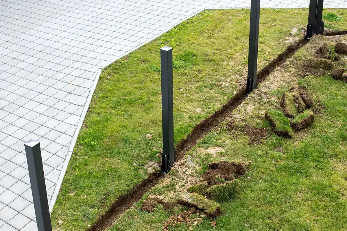 Vorbereitung für den **Zaun selber bauen**: Pfosten sind gesetzt, ein Graben für das Fundament ist ausgehoben, Rasenstücke liegen daneben.