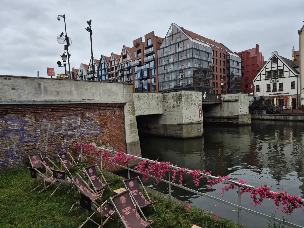 Nad rzeką, pod zielonym mostem, stoją leżaki. W tle nowoczesne budynki i zabytkowa kamienica.