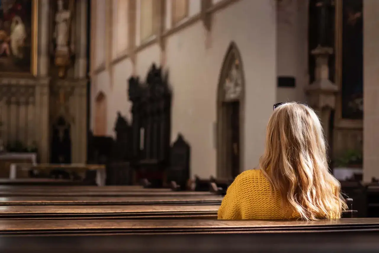 Frau mit blonden Haaren in gelbem Pullover sitzt in einer Kirche. Ein stilles Baptisten Frauenbild der Besinnung.