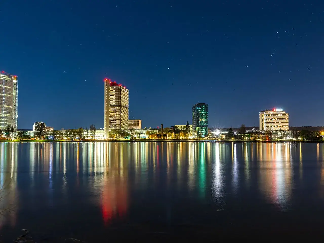 Bonn bei Nacht: Hochhäuser spiegeln sich im Wasser. Entdecke die bonn geheimtipps, die diese Skyline zu etwas Besonderem machen.