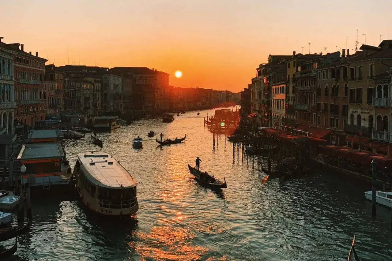 Tramonto sul Canal Grande, Venezia. Le fondamenta della città si specchiano nell'acqua dorata, con gondole e vaporetti che navigano al calar del sole.