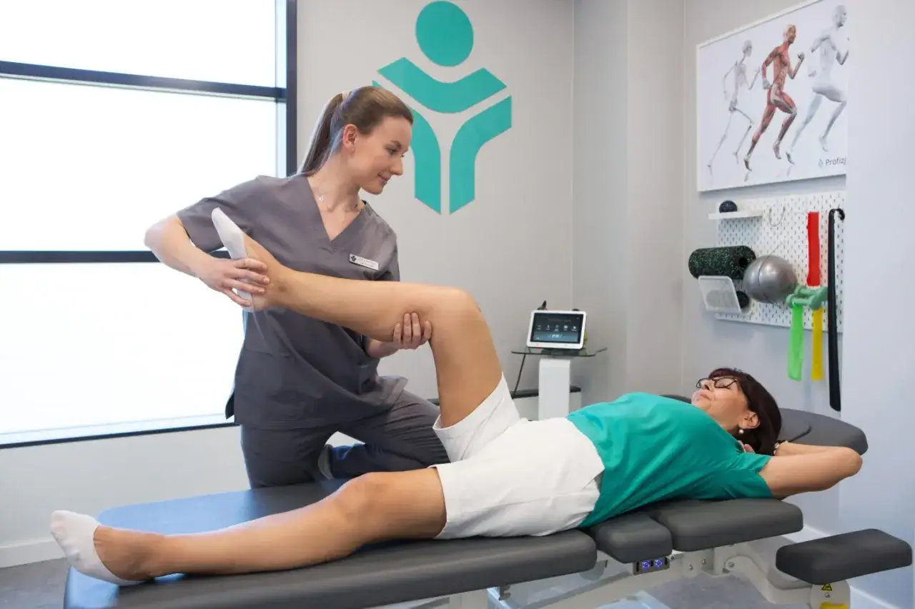 Fizjoterapeuta wykonuje ćwiczenia z pacjentką, która leży na kozetce. W tle logo kliniki i sprzęt rehabilitacyjny.