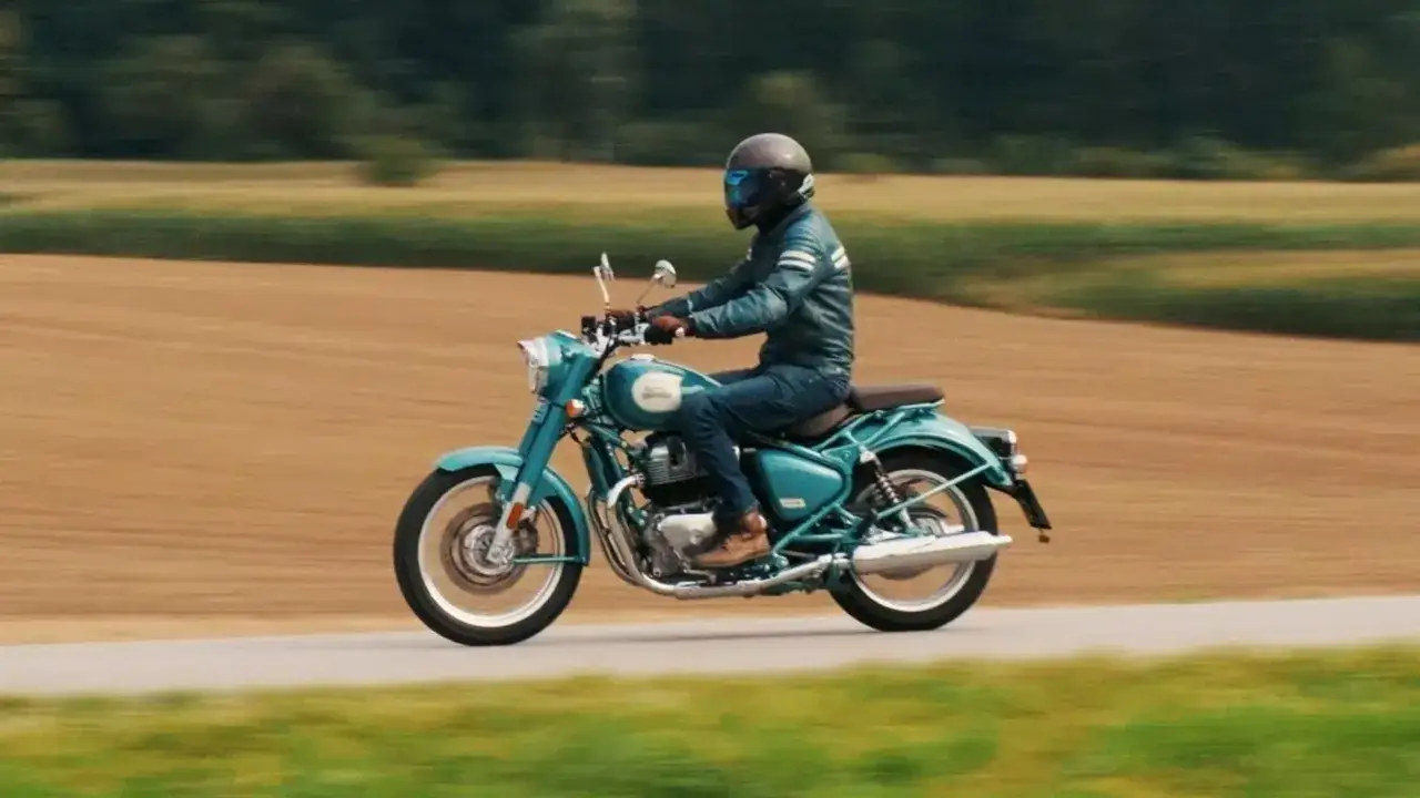 Mężczyzna w kasku i skórzanej kurtce jedzie na turkusowym motocyklu Royal Enfield Classic 650 przez wiejskie krajobrazy.