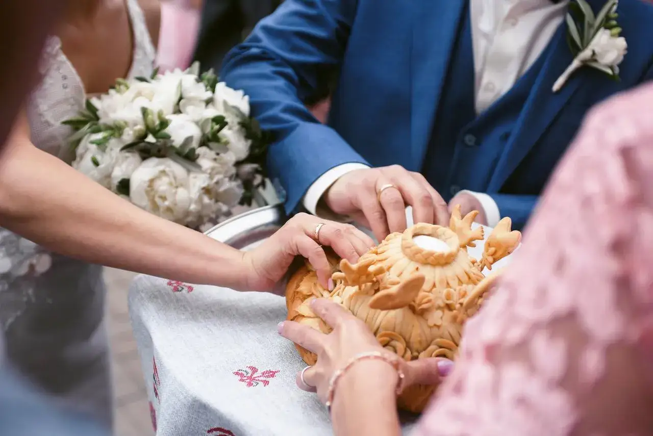 Manos de novios tocando un pan decorado, parte de rituales de boda en diferentes culturas. Un ramo de peon&iacute;as blancas adorna la escena.