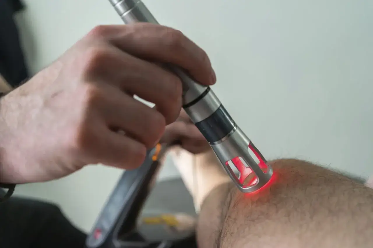 Terapia laserem rehabilitacja: dłoń terapeuty trzyma urządzenie emitujące czerwone światło na skórę pacjenta.