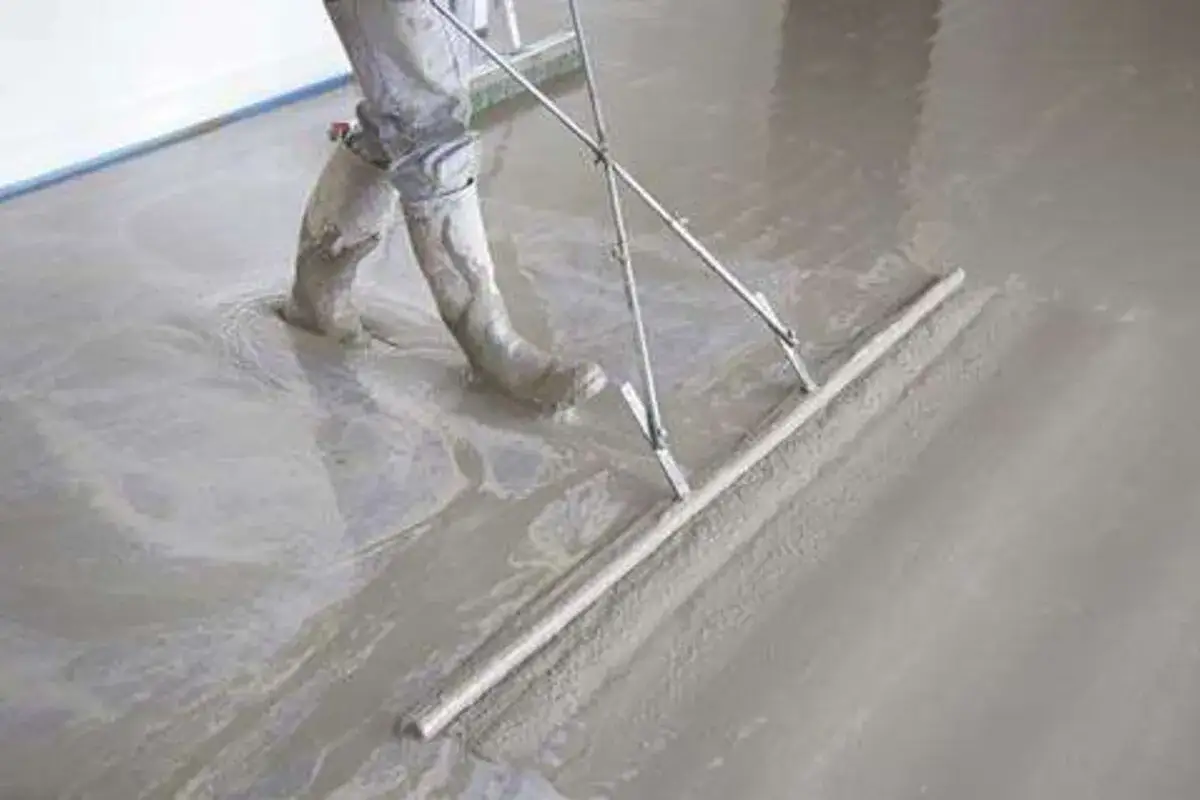 Un ouvrier en bottes travaille sur une chape fra&icirc;chement coul&eacute;e, utilisant une r&egrave;gle de ma&ccedil;on pour lisser le b&eacute;ton.