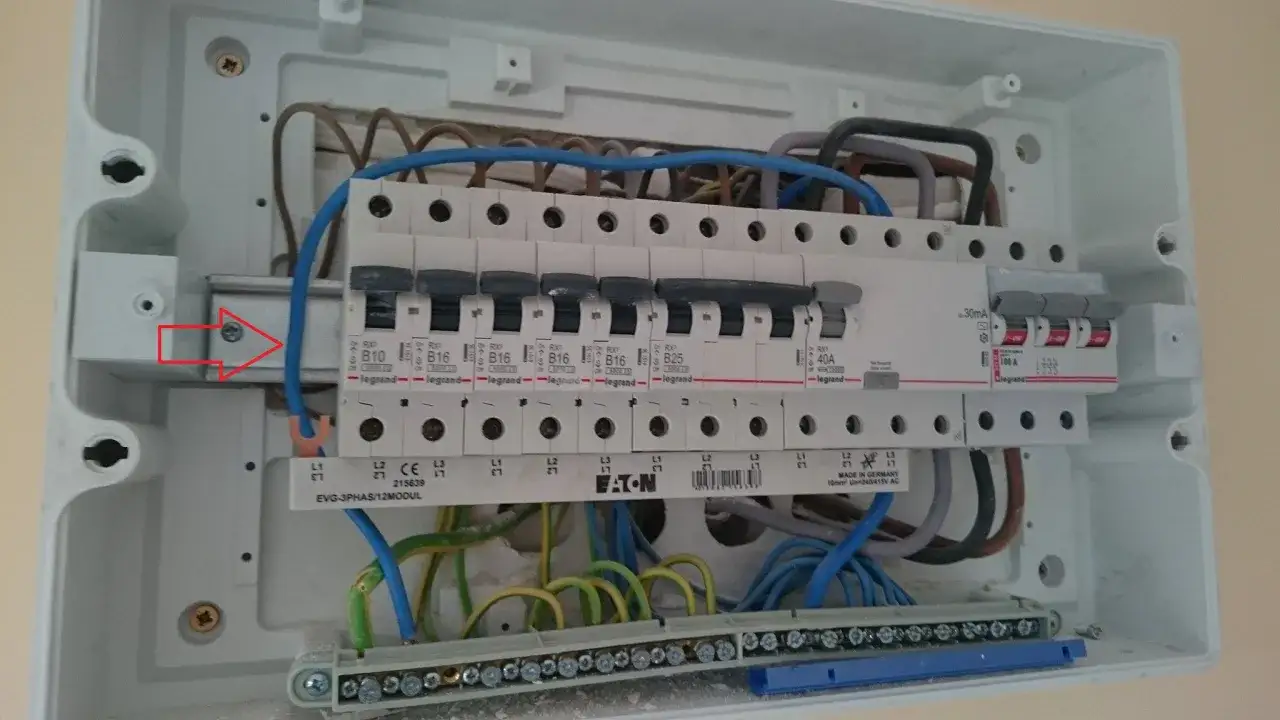 W skrzynce elektrycznej widać zabezpieczenie przedlicznikowe, bezpieczniki i przewody. Czerwona strzałka wskazuje śrubę mocującą szynę.