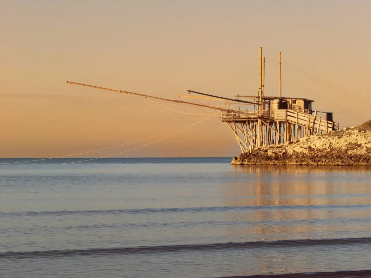 La chianca vieste, un trabucco di legno su scogli, si specchia nel mare calmo al tramonto.