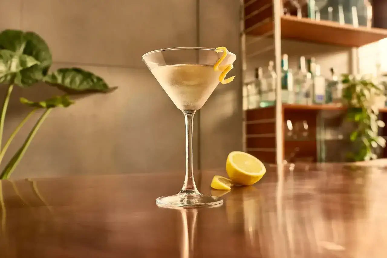Elegancki martini wstrząśnięte nie mieszane, ozdobione skórką cytryny, stoi obok połówki cytryny na błyszczącym blacie.