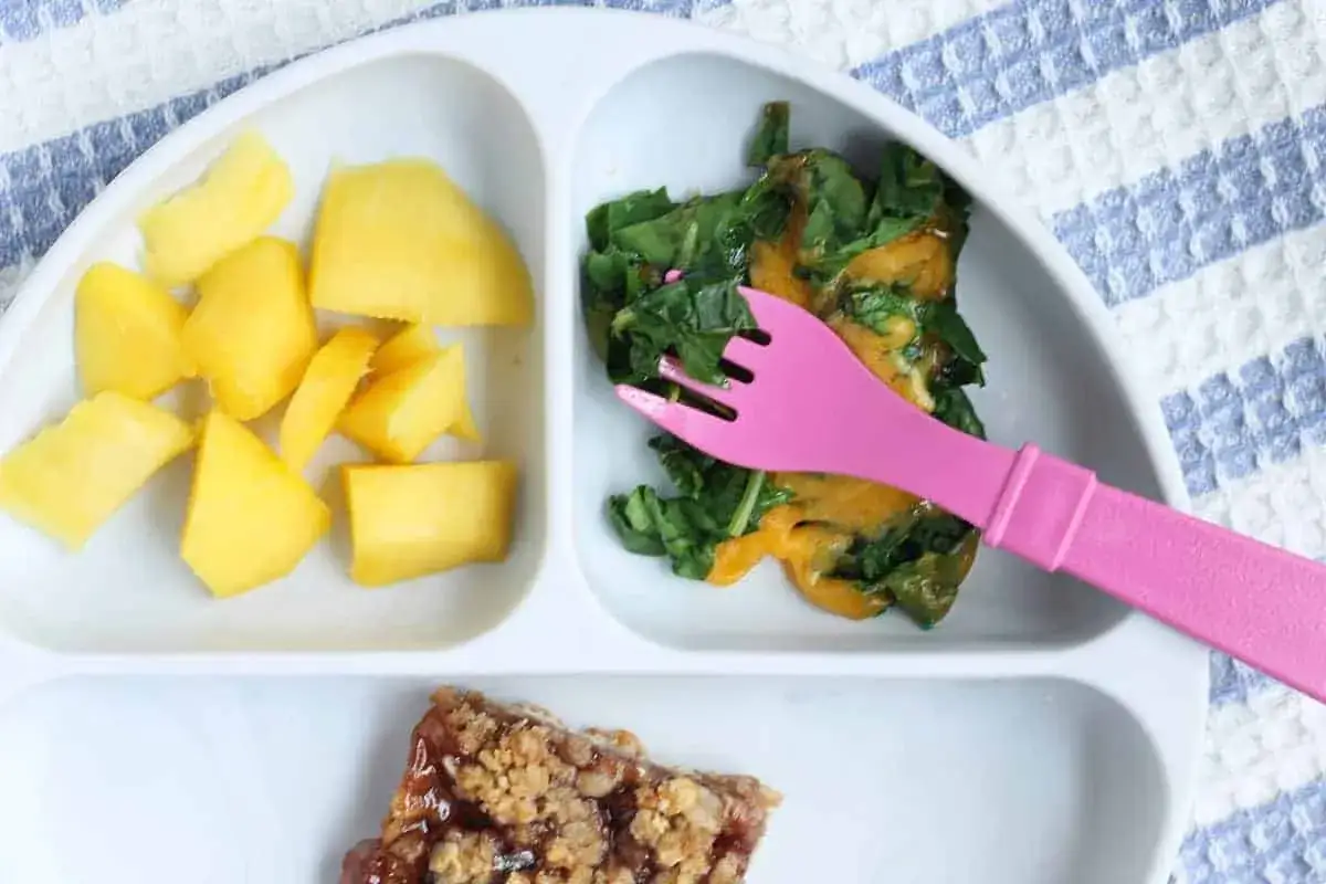 Plato dividido con trozos de mango, espinacas con queso y un trozo de barrita de cereales. ¡Ideas de recetas con verduras para niños!