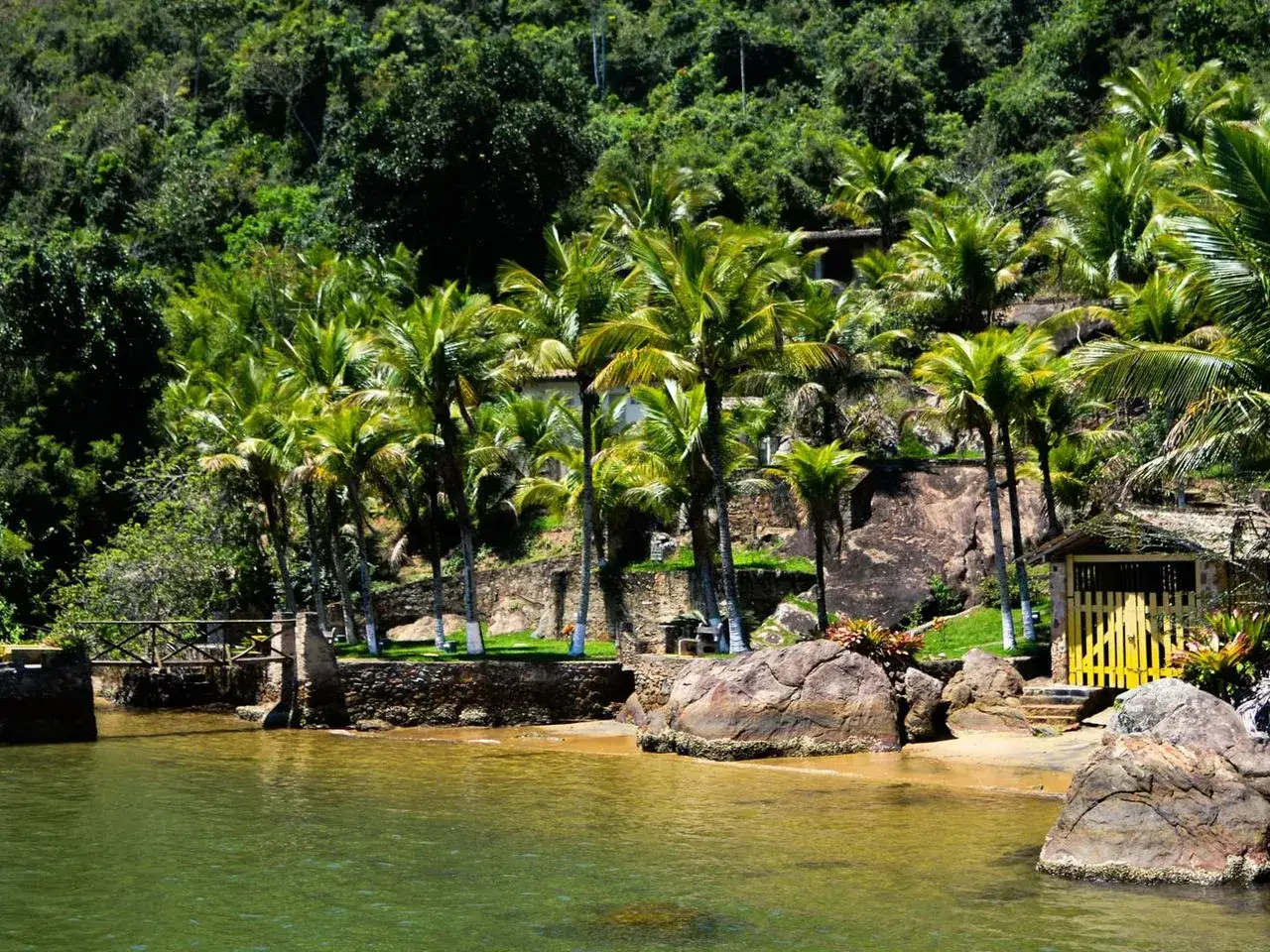 Paraty, Brasilien: Tropischer Strand mit Palmen, Felsen und einem gelben Torhaus.