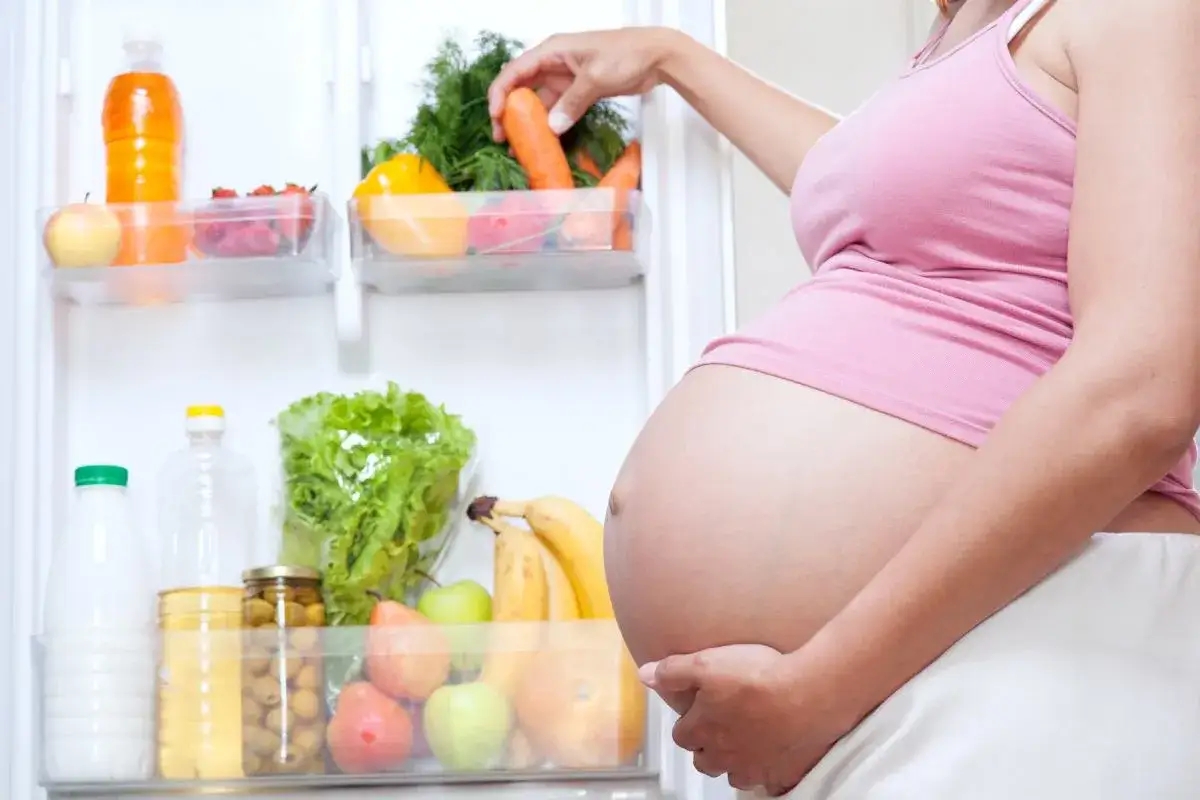 Une femme enceinte choisit une carotte dans son r&eacute;frig&eacute;rateur rempli de fruits, l&eacute;gumes et eau.