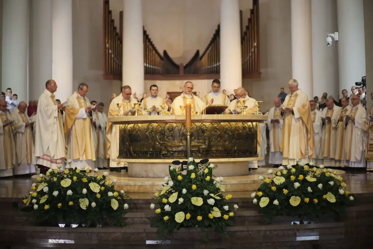 Uroczysta msza w kościele, kapłani w szatach liturgicznych przy ołtarzu. W tle organy, przed ołtarzem bukiety kwiatów.