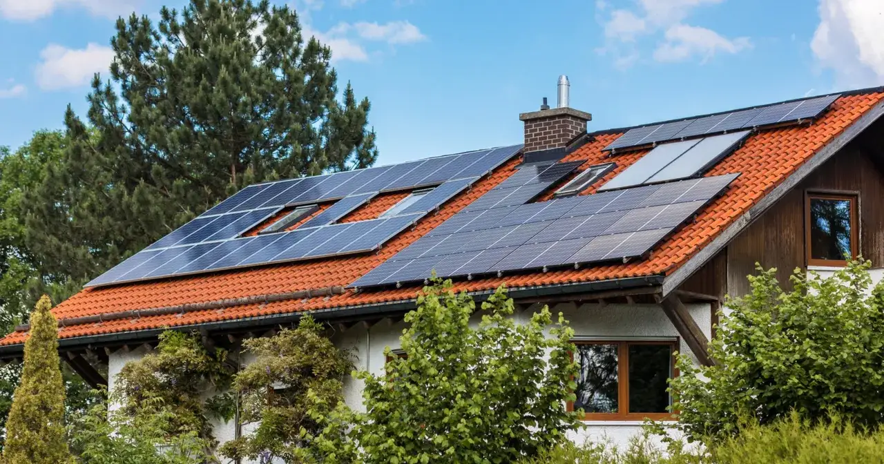 Ein **autarkes Haus** mit vielen Solarmodulen auf dem roten Ziegeldach, umgeben von gr&uuml;nen B&auml;umen und blauem Himmel.