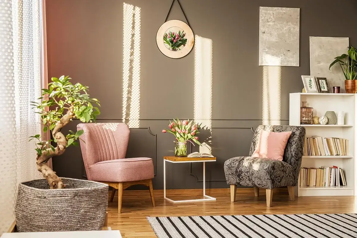Un angolo relax con due poltrone, un tavolino con fiori e libri, e una libreria piena. La luce del sole crea un'atmosfera accogliente.