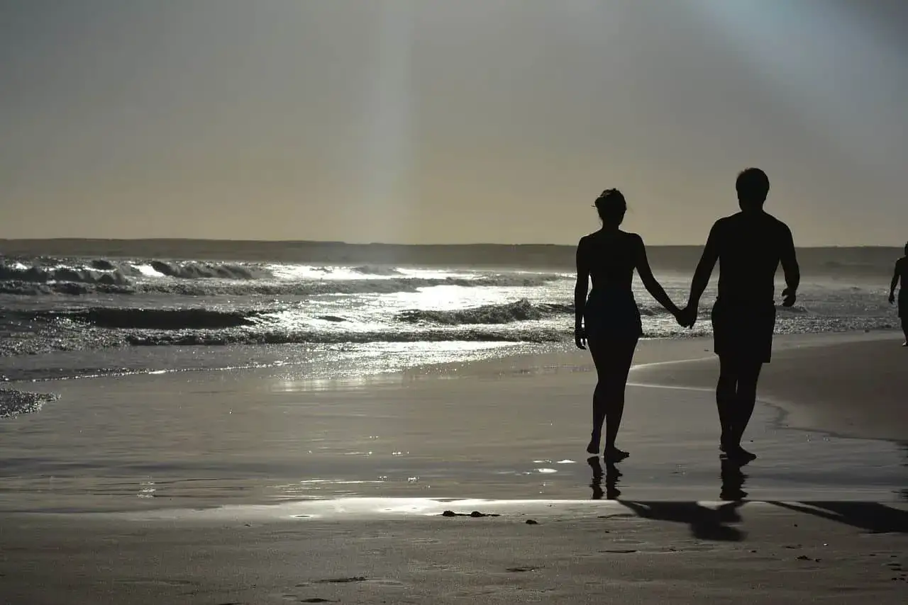 Silhouettes d'un couple main dans la main sur une plage au coucher du soleil. Pourquoi un homme cache qu'il a une copine ? Peut-être pour préserver ce moment de quiétude.