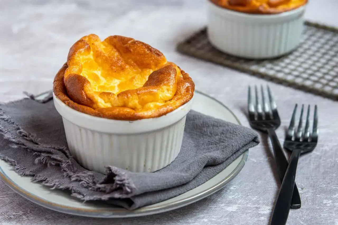 Un suprême soufflé, doré et aérien, servi dans un ramequin blanc sur une assiette grise, prêt à être dégusté comme accompagnement parfait.
