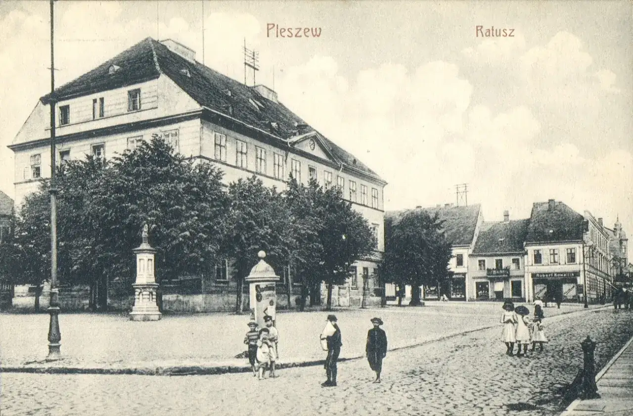 Rynek w Pleszewie Wielkopolskim z ratuszem i pomnikiem. Dzieci bawią się na brukowanym placu.