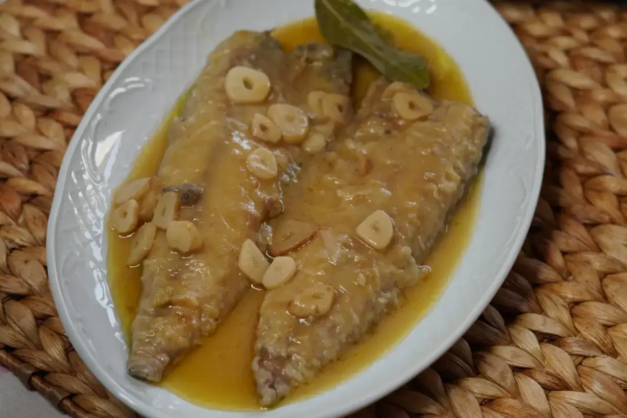 Dos filetes de caballa en escabeche, cubiertos de rodajas de ajo y una hoja de laurel, servidos en un plato blanco sobre una estera tejida.